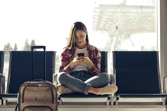 Air travel concept with young casual girl sitting with hand luggage suitcase. Airport woman on smart phone at gate waiting in terminal.