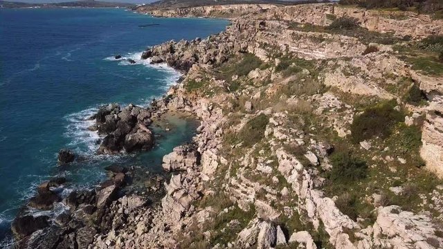 AERIAL: Rocky Cliff beach with green grass blue water. Flyover shot with camera tilting down. Mellieha, Malta