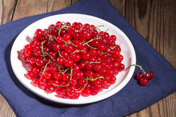 Fresh red currants in a white plate on a natural brown wooden background on line blue napkin. Close-up. Vegetarianism