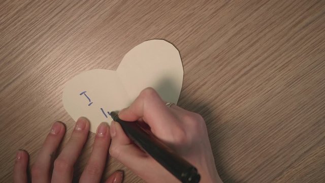 Hand's of young girl write phrase i luv u on white paper heart. Romantic stuff. Close up. Up view.