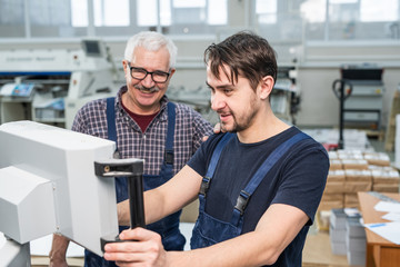 Smiling young bearded intern of printing factory operating machine under control of experienced...