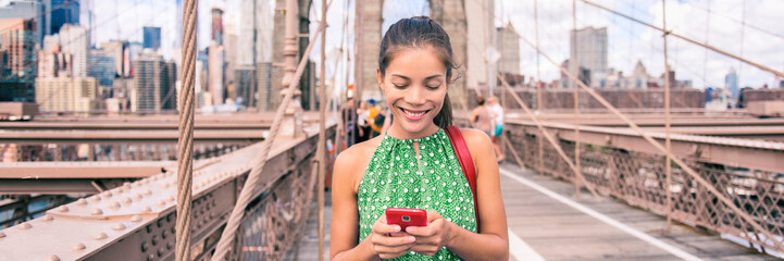 Happy woman using phone walking in NYC on Brooklyn Bridge. New York city lifestyle young Asian girl...