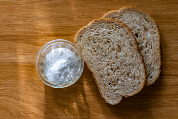 Two pieces of black bran fresh bread with white salt in a salt shaker, standing on a wooden brown table in the sunlight.