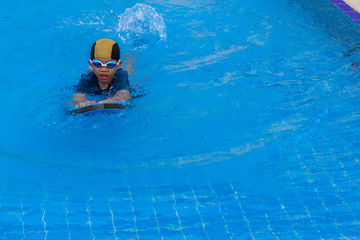 6 years old young boy a swimmer wear goggle with the dark blue suit and yellow striped black cap warm up with swimming kickboard before praticing to swim in the child blue swimming pools.