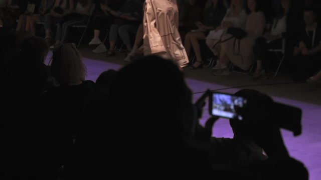 fashion show of brand new collection of modern clothing, slender unrecognizable model walks along purple path, viewers shoot videos on mobile devices and phones