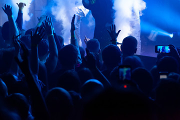 Hand with a smartphone records live music festival, Taking photo of concert stage. Party at nightclub with crowd of people raising hands with phones.