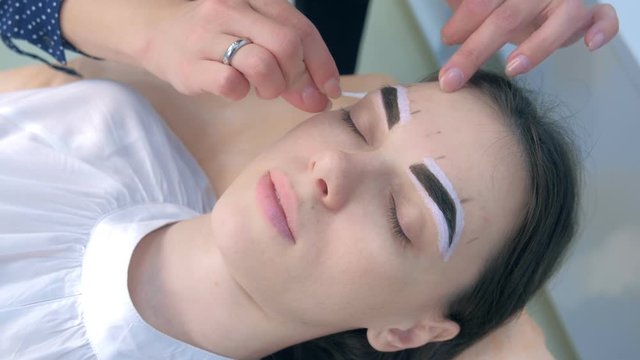 Beautician wiping white paint from eyebrows during tinting procedure, hands closeup. Cosmetologist is making tint brows. Dark brown natural henna paint on woman eyebrows, face closeup.