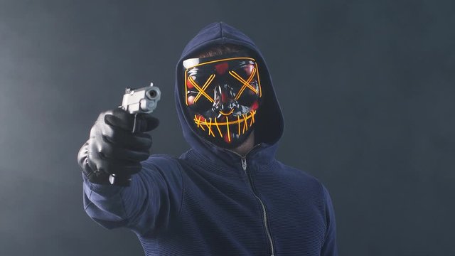 Young incognito man in mask stand with gun isolated over smoky space. Anonymous burglary male look at camera, engaged in cyberattack, in crime.