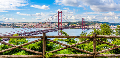 Fototapeta premium Landscape of the 25th of April bridge over the Tagus river estuary into Lisbon skyline as seen from the hills of Almada with dramatic clouded sky empty for text in Lisbon, Portugal