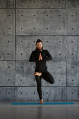 Fototapeta na wymiar A young bearded man in black sportswear is practicing yoga against a gray wall.