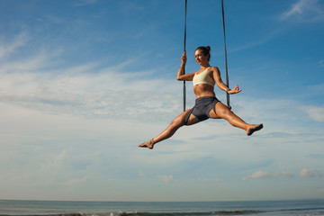 aerial yoga meditation beach workout - young attractive and healthy woman practicing aero-yoga training balance body and mind control hanging from ropes above the sea