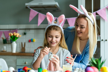 Funny, cheerful, joyful mum teaching, training her cute, pretty, small, little daughter draw, paint, decorate easter eggs, together wearing bunny ears, preparing for Easter