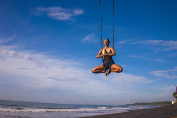 aerial yoga meditation beach workout - young attractive and healthy woman practicing aero-yoga training balance body and mind control hanging from ropes above the sea
