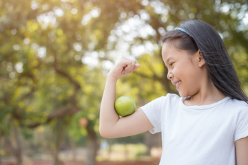 photo happy Little asian girl child standing with big smile. girl with green apple showing biceps..fresh healthy green bio background with abstract blurred foliage and bright summer sunlight