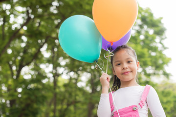 happy little asian girl child playing with colorful toy balloons outdoors. trees and green gardens...