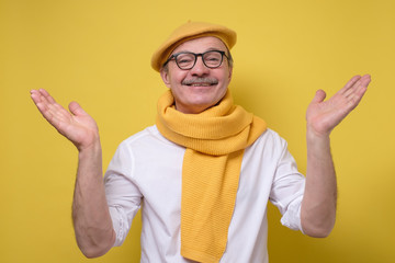 Hispanic senior man in glasses and yellow beret and scarf spreading hands up welcoming his friend looking with a smile at camera. Studio shot. Positive facial human emotion.