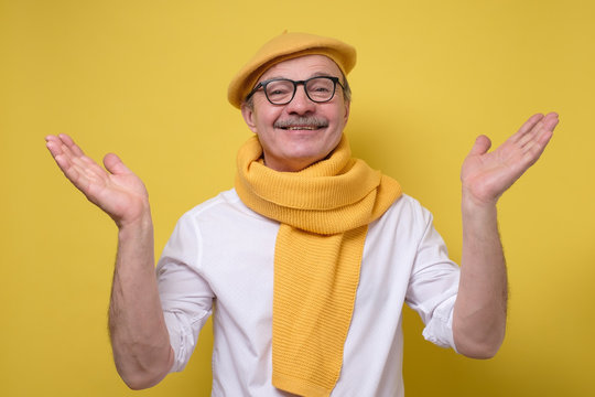 Hispanic Senior Man In Glasses And Yellow Beret And Scarf Spreading Hands Up Welcoming His Friend Looking With A Smile At Camera. Studio Shot. Positive Facial Human Emotion.