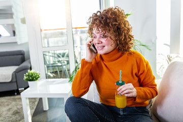 Happy cheerful young woman talking on the phone at home, smiling teen girl answering call by cellphone sitting on sofa, having pleasant funny conversation speaking by mobile and drinking orange juice