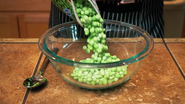 Cook pours frozen peas from plastic packaging into glass bowl, slowmo