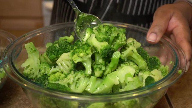 Cook stirs steamed broccoli in glass bowl with tablespoon, slow motion