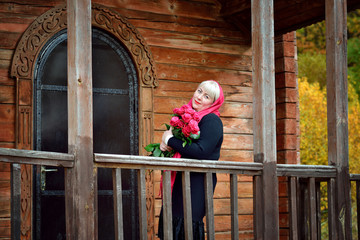 A plump middle-aged woman stands on the porch of an old wooden house in a red scarf and black dress, looking at the camera and smiling with red roses in her hands.