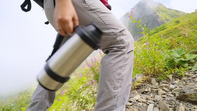 Woman backpacker hiking on rocky path in sunny day. close-up view legs in trekking boots and thermo flack in hands. Tourist climbing on mountain summit, camera following faceless female goes uphill. 