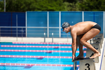 Young muscular swimmer in low position ready to jump on starting block in a swimming pool.