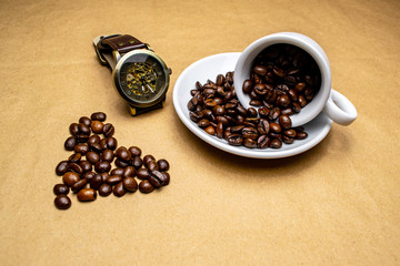 Heart made of coffee, a watch and a fallen white cup with coffee beans on a beige background