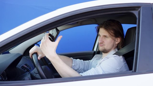 Brunette driver is taking photos while driving a car. He is sitting in white car with chroma key background. Young guy in casual clothes is making duckface and taking selfie.