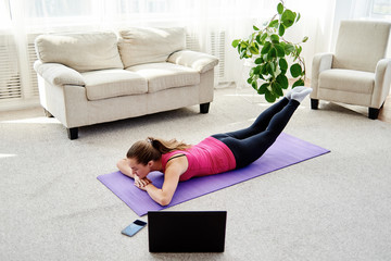 Beautiful young woman doing exercise for back on floor at home, online training on laptop computer, copy space. Full length portrait. Yoga, pilates, working out exercising