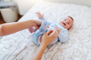 Close up of caring caucasian mother putting tiny socks on baby's feet. Baby lying on bed. Selective focus on foot.