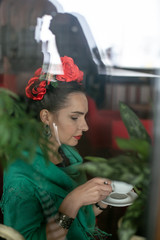 vertical photo of a young woman in bright clothes and flowers in her hair who is sitting with a cup of coffee in her hands