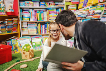 Caring father sitting on the floor with his daughter on the floor in bookstore and reading stories to her. All around are shelves with books for children.