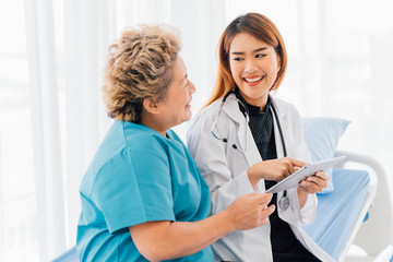 Young Asian female doctor in uniform laughing while giving advice about treatment with digital tablet to senior female patient and sitting on bead in hospital ward