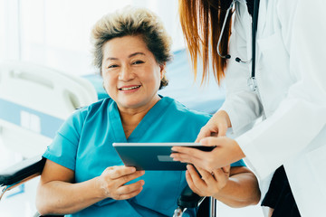 From above of satisfied Asian aged female patient on wheelchair smiling and looking at camera while checking medical history with doctor on digital tablet