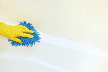 A female hand in a yellow rubber glove with a blue mop cloth washes and polishes the surface of a white bathtub. Spring, regular spring cleaning. Bathroom Cleaning Concept. Copy space for text.