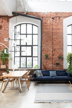 Loft Apartment In Industrial Style With Big Window. Interior Of Living Room With Design Navy Sofa And Stylish Table With Chairs. Bright Indoors.