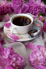 White cup or mug with hot black tea or coffee with chocolate candy and flowers petals on a saucer surrounded by fresh pink peonies, wonderful female morning or a coffee break