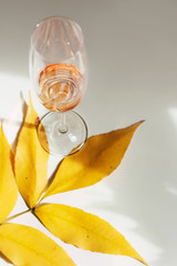 Glass of pink wine or fruit juice without pulp stands on a branch with bright yellow leaves. Simple minimalistic still life with an alcoholic drink on a white table. Sunny photo with shadows