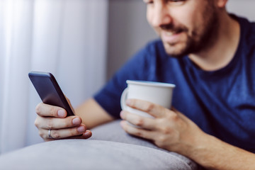 Close up of attractive smiling unshaven man leaning on sofa, holding mug with coffee and replying on messages on social media.