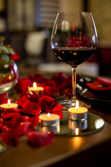 vertical photo of a glass with wine against the background of candles and rose petals