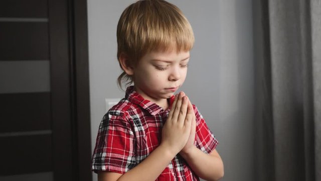 A blond boy prays alone in his room. The child holds his hands together.