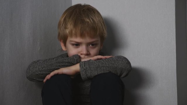 A scared, lonely blond boy sits on the floor in the corner of the room.