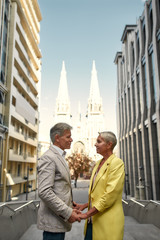 Always together. Elegant and stylish mature couple holding hands and looking at each other with smile while standing against beautiful catholic church