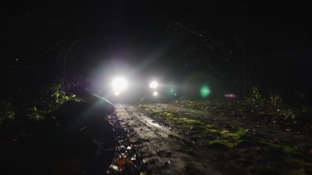 A frightened girl quickly runs away from car persecution her along a country road at night. Woman stumbling falls to the ground. Silhouette of a running girl in the headlights of a car
