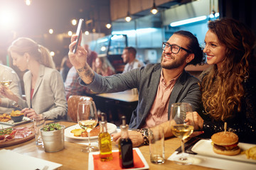 Cute caucasian fashionable couple sitting in restaurant at dinner and taking selfie.