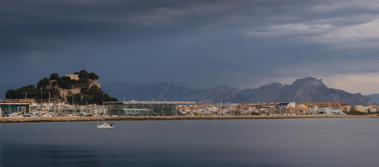 A sea-side panoramic view of the Spanish port city of Denia with dark clouds in the sky. The skyline shows the harbor and the castle on the left. A sailboat is anchored in the foreground.