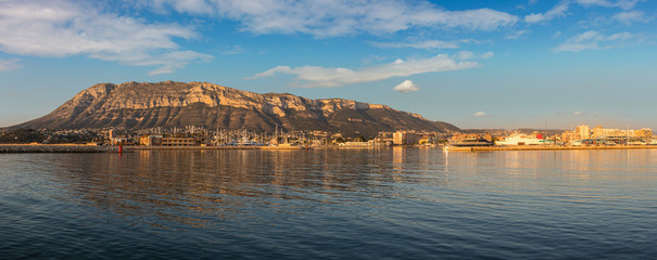 A sea-side panoramic view of the Spanish port city of Denia shortly after sunrise. The skyline and the Montgo mountain are illuminated by the warm morning light. Ships are in the harbor.