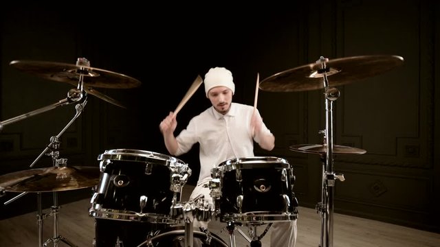 Handsome guy in white actively plays on drum kit in on black background. Closeup
