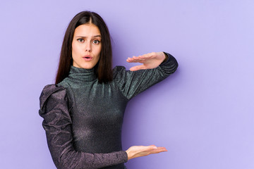 Young caucasian woman isolated on purple background holding something with both hands, product presentation.